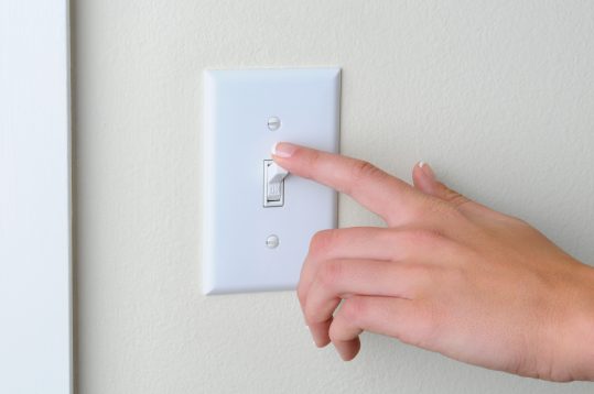 Womans hand with finger on light switch, about to turn off the lights. Closeup of hand and switch only. Horizontal format.